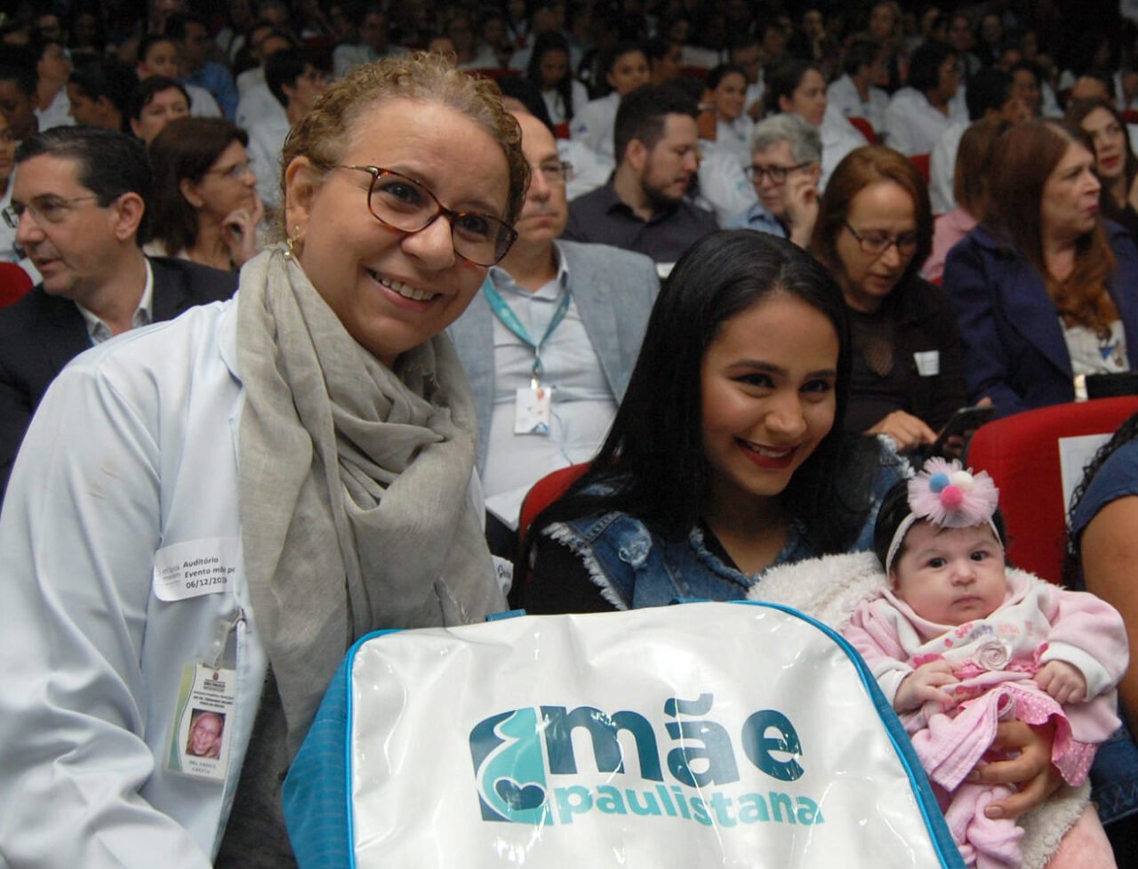 Dra Greice Chetta al lado de uma mamãe com seu bebê no colo, durante um evento