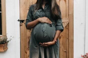 O Mistério da Bolsa das Águas e Seu Rompimento - imagem de mulher grávida com as mãos acima e abaixo da barriga, em vestido verde com porta de madeira atrás